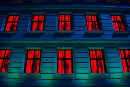 Foto de Escena nocturna cautivadora con una fachada urbana de rascacielos bañada en azul con ventanas rojas sorprendentemente iluminadas, creando un cuadro urbano abstracto. - Imagen libre de derechos