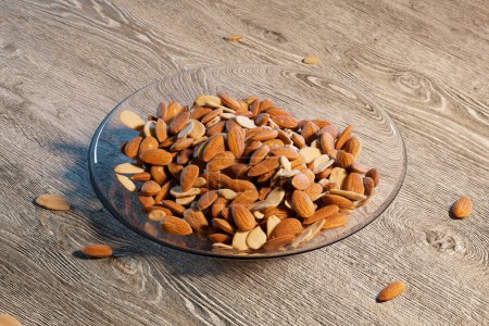Top view of an abundant glass bowl overflowing with nutritious, unprocessed almonds, set against the rich texture of a dark wooden surface ideal for food themes.