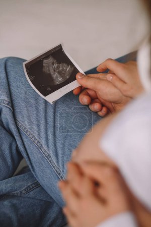 Close up photo of pregnant woman and her husband holding their hands on  pregnant belly and looking at the ultrasound photo. Love concept. Family concept