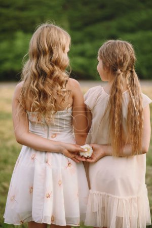 Rare view of two little girls with long blonde hair in summer dresses holding a rose flower in their hands behind their backs on nature background. The concept of childhood friendship. Friends