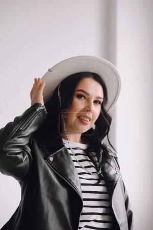 Portrait of smiling brunette woman in white hat and black leather jacket indoor. Beauty and fashion. Copy space for text. Good for wallpaper or print magazine