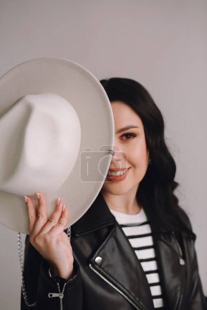 Smiling brunette woman in black leather jacket holding a white hat in front of the face on white background. Copy space for text. Good for wallpaper or print magazine