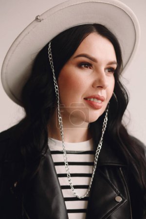 Portrait of beautiful brunette woman in white hat and black leather jacket in the studio. Beauty and fashion. Smiling face. Good for wallpaper or print magazine