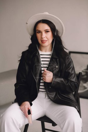Beautiful brunette makeup artist in white trousers, black leather jacket and white hat sitting on the chair and holding tonal cream in her hand. Beauty and fashion