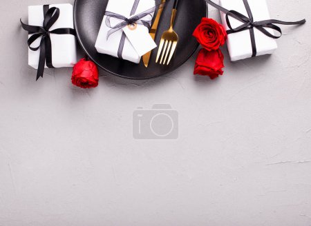 Téléchargez les photos : Bordure de table romantique. Assiette noire, couteau, fourchette, boîtes avec cadeaux et fleurs de roses rouges sur fond gris texturé. Vue de dessus. Place fr texte. Carte postale romantique. - en image libre de droit