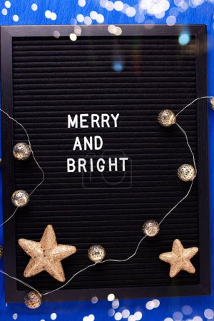 Photo for Christmas postcard. Letterboard with phrase merry and bright and golden decorations  on bright blue paper textured background. Top iew. Bokeh light. - Royalty Free Image
