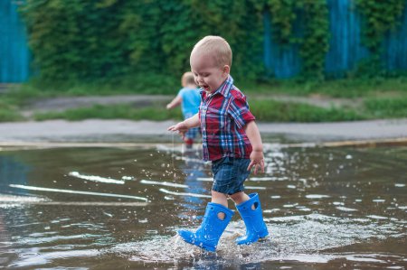 Téléchargez les photos : Joyeux tout-petit garçon courant à travers les flaques dans des bottes en caoutchouc. Des éclaboussures d'enfants après la pluie dehors - en image libre de droit