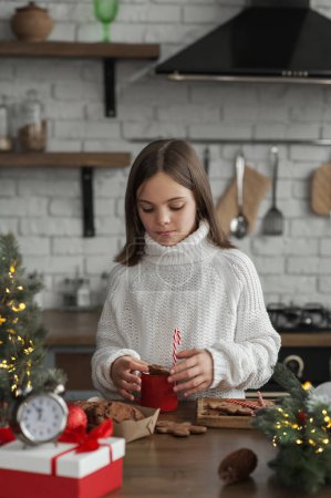 Téléchargez les photos : Fille en chapeau Santa pour Noël mange des biscuits et du cacao à la maison dans la cuisine. - en image libre de droit