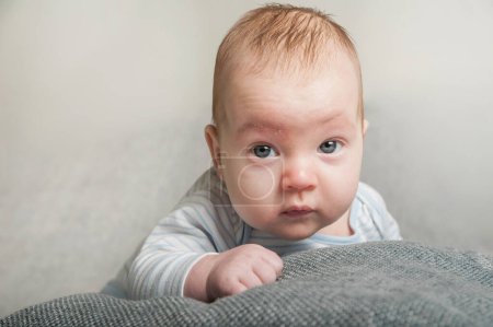 Ein Baby mit sanftem Gesichtsausdruck liegt bequem auf einer strukturierten Decke und blickt neugierig in die Kamera. Die sanfte Beleuchtung unterstreicht die beruhigende Atmosphäre und hebt die Babys hervor