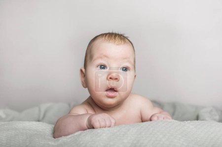 Téléchargez les photos : Un bébé avec une expression douce se trouve confortablement sur une couverture texturée, regardant vers la caméra avec une curiosité aux yeux larges. La lumière douce améliore l'atmosphère apaisante, mettant en valeur les bébés - en image libre de droit