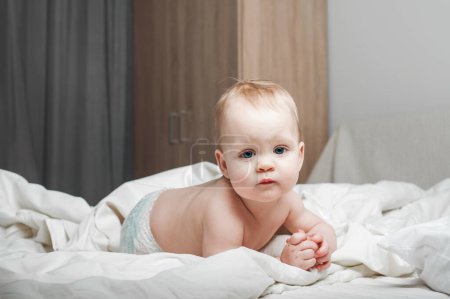 Téléchargez les photos : Portrait de beau bébé de 11 mois en couche sur le lit gros plan. Soins de la peau, concept de santé pour les enfants. Dentition, mains sur la bouche des tout-petits. - en image libre de droit