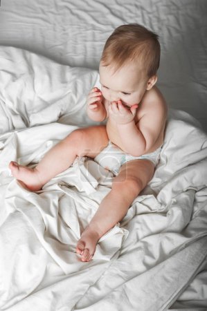 Téléchargez les photos : Portrait de beau bébé de 11 mois en couche sur le lit gros plan. Soins de la peau, concept de santé pour les enfants. Dentition, mains sur la bouche des tout-petits. - en image libre de droit