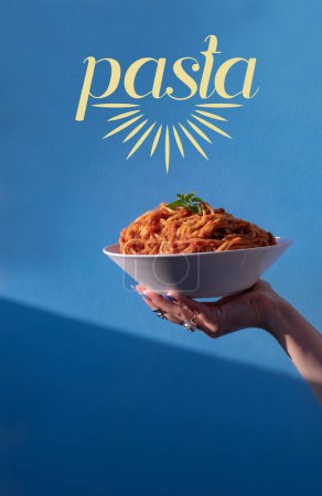 Téléchargez les photos : Main féminine tenant une assiette de pâtes spaghetti avec sauce tomate dans une assiette blanche sur fond bleu avec le mot pâtes. Le concept de la cuisine italienne moderne. Gros plan focus sélectif - en image libre de droit