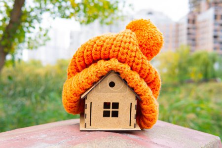 Chunky knitted hat put on a wooden house model placed outdoors with a city background. Roof sealing maintenance and renovation concept.