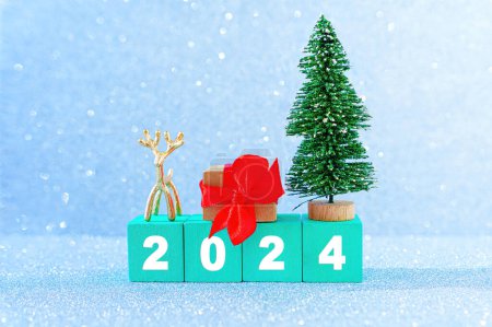 Neujahr 2024 Hintergrund aus grünen Buchstabenblöcken aus Holz, Spielzeug-Weihnachtsbaum, winzige Hirschfigur und verzierte Geschenkschachtel vor glitzerndem Hintergrund, der Schnee imitiert.