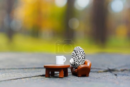 Photo for Miniature human brain figurine drinks coffee sitting in a armchair in the woods. Creative relax and city break concept. - Royalty Free Image