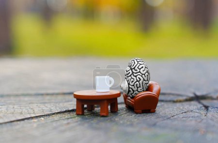 Photo for Miniature steel copy of a human brain drinks coffee sitting in a armchair outdoors. Creative city escape and mind relaxation concept. - Royalty Free Image