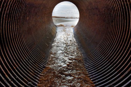 Foto de Vista desde el interior de un gran tubo de drenaje oxidado que conduce al océano, con un arroyo de agua y arena que fluye río abajo hacia la playa. - Imagen libre de derechos