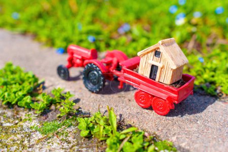 Téléchargez les photos : Petite cabane en bois dans la remorque d'un tracteur jouet, placée dans un beau jardin par une journée ensoleillée. Concept de livraison cabine jardin. - en image libre de droit