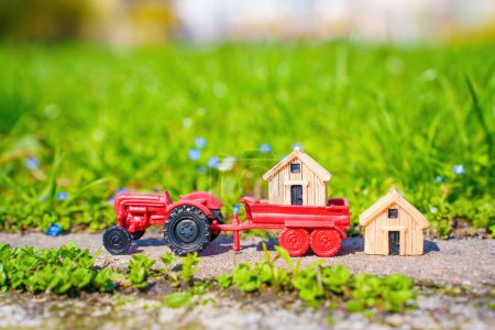 Téléchargez les photos : Tracteur jouet rouge et deux petites maisons en bois sur une route pavée dans le jardin contre une pelouse verte luxuriante. Concept d'approvisionnement et de service des hangars de jardin. - en image libre de droit