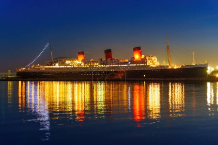 Téléchargez les photos : Vue imprenable sur le navire emblématique Queen Mary alors qu'il illumine le ciel nocturne, jetant un beau reflet sur l'eau ci-dessous. Capturé à Long Beach, Californie. - en image libre de droit