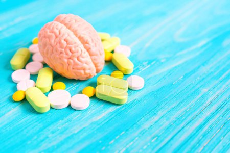 Modèle détaillé d'un cerveau humain entouré de pilules ovales et rondes de couleur blanche, verte et jaune, placées sur un fond en bois bleu avec espace de copie. Concept de conscience piégée.