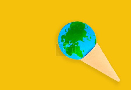 Téléchargez les photos : Concept de réchauffement et de changement climatique : globe miniature niché dans un délicieux cône de gaufre à la crème glacée isolé sur fond jaune avec espace de copie. - en image libre de droit
