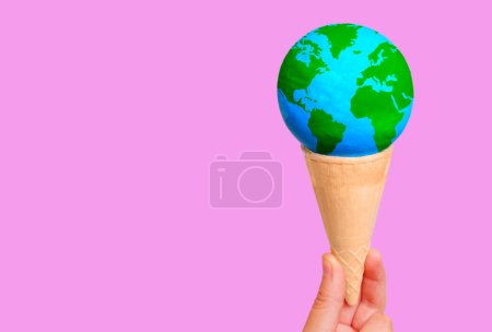 Téléchargez les photos : Main tenant un globe miniature délicatement placé à l'intérieur d'un cône de gaufre croustillant sur fond rose avec espace de copie. Désir créatif et concept lié à l'exploration mondiale. - en image libre de droit