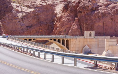 Visiteurs traversant le pont en béton qui enjambe le barrage Hoover, face à une montagne robuste, rouge-brun.