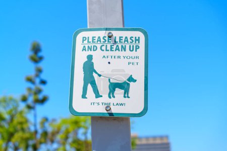 Rectangular sign posted on a pole in a grassy area, instructing pet owners to leash their dogs and clean up after them.