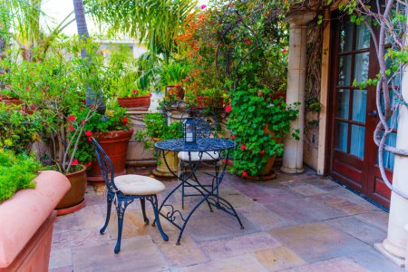 Encantador patio exterior con una mesa redonda de hierro forjado y dos sillas, rodeado de exuberante vegetación y plantas florecientes.