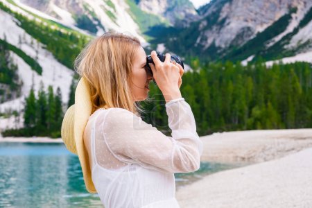 Foto de Joven fotógrafa sosteniendo una cámara y tomando fotos de los Alpes. Una chica con un vestido blanco y sombrero disfrutando de la vista de las montañas. - Imagen libre de derechos
