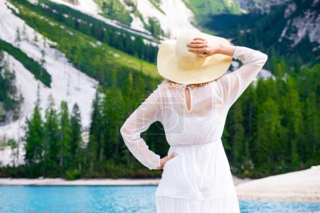 Foto de Joven hermosa mujer en un vestido blanco sosteniendo un sombrero en la cabeza contra el lago Braies y las montañas. - Imagen libre de derechos