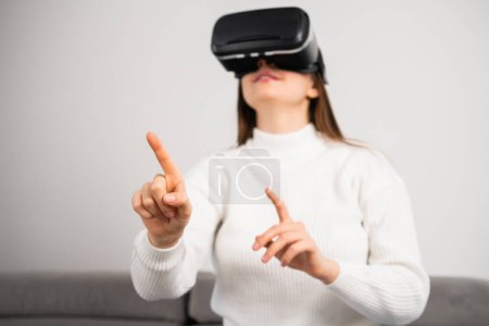 Foto de Curious housewife in VR goggles swipes virtual interface to choose game. Young woman rests on couch tasting modern innovations at home - Imagen libre de derechos