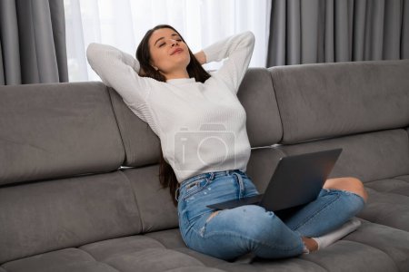 Photo for Relaxed young woman lounges on a comfortable sofa, arms crossed behind her head and holding laptop on her knees in a cozy modern living room. - Royalty Free Image