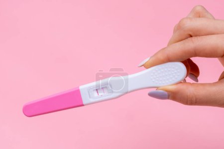 Womans hand holding a positive pregnancy test with two stripes on pink background with copy space.