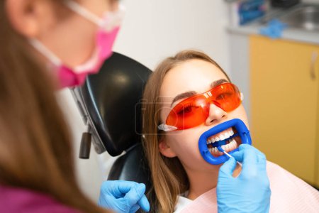 Dentist doing a whitening teeth procedure for young blond woman at the clinic.