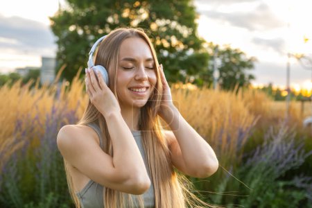 Téléchargez les photos : Joyeux jeune femme aime écouter de la musique et chanter tout en étant assis dans le parc au coucher du soleil. Dame heureuse dans les écouteurs repose passer du temps dans la nature - en image libre de droit