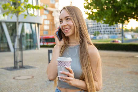 Foto de Mujer joven feliz con el pelo largo y suelto rubia pasa tiempo en el parque hablando con el novio por teléfono. Lady se ríe alegremente bebiendo café al aire libre - Imagen libre de derechos