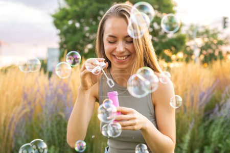 Foto de Mujer joven feliz con el pelo largo suelto sopla burbujas de jabón riendo en el jardín de la ciudad. Sonriente dama sopla burbujas divirtiéndose en el parque por la noche - Imagen libre de derechos