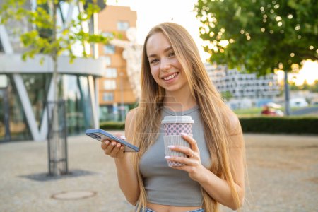 Foto de Joven mujer de pelo rubio sonriente revisando aplicaciones de teléfono celular y bebe café de pie en la calle - Imagen libre de derechos
