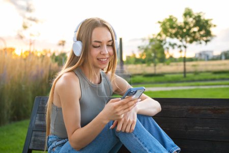 Foto de Mujer rubia despreocupada escuchando música usando auriculares y seleccionando música a través de un teléfono inteligente sentado en el banco en el parque. - Imagen libre de derechos