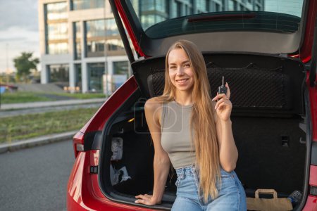 Foto de Retrato de mujer rubia joven con el pelo largo sentado en el maletero del coche rojo nuevo y con las llaves. - Imagen libre de derechos