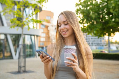Foto de Atractivo cabello rubio mujer utiliza su teléfono inteligente y bebe café en la calle. - Imagen libre de derechos