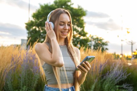 Foto de Retrato de una joven rubia escuchando música con auriculares y sonriente composición selectiva en el parque - Imagen libre de derechos