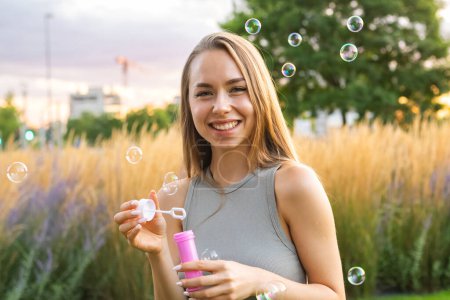 Foto de Una joven alegre con el pelo largo y fluido crea burbujas de jabón al atardecer en el parque. - Imagen libre de derechos