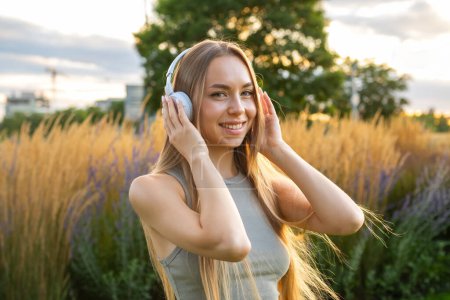 Foto de Mujer joven rubia escuchando música usando auriculares mientras descansa en el parque. - Imagen libre de derechos