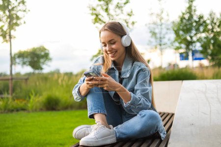 Foto de Una chica de pelo rubio está sentada en un banco del parque con las piernas cruzadas, dando la bienvenida al sol de la noche y escuchando música a través de auriculares. - Imagen libre de derechos