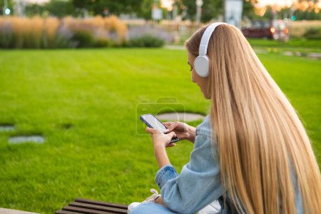 Foto de Chica rubia se sienta con su espalda en los auriculares y utiliza un teléfono móvil en un claro en el parque. - Imagen libre de derechos
