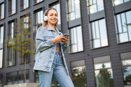 Foto de Joven mujer rubia en denim en auriculares escucha música en la calle. - Imagen libre de derechos
